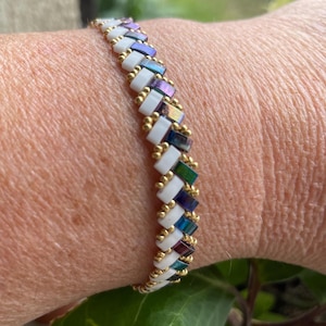 Peut inclure: Un bracelet de perles avec un design blanc, doré et irisé. Le bracelet présente des perles rectangulaires blanches, des perles rectangulaires irisées dans les tons violets, bleus et verts, et de petites perles dorées. Le bracelet est porté au poignet.