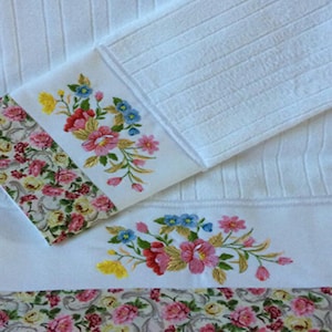 May include: Two white towels with embroidered floral designs in pink, yellow, blue, and green. The towels have a floral patterned border.