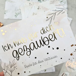 May include: A white card with gold foil lettering that reads 'Ich hab dich  gezaubert!' and 'Danke für dein Vertrauen.' The card has a watercolor background with black and gold dots and a black and white floral design.
