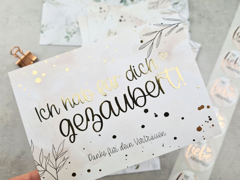 May include: A white card with gold foil lettering that reads 'Ich hab dich  gezaubert!' and 'Danke für dein Vertrauen.' The card has a watercolor background with black and gold dots and a black and white floral design.