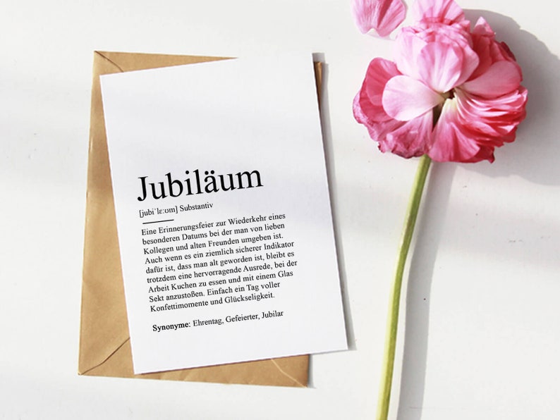 May include: A white card with the word "Jubil&auml;um" in black text. The card defines the word as a celebration of a special date, surrounded by loved ones and friends. The card is on a brown envelope with a pink flower on a white background.
