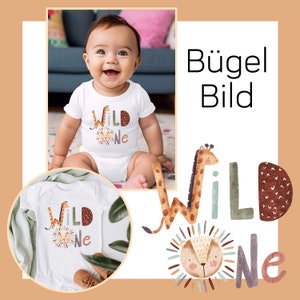 Pode incluir: Um macacão branco com um gráfico "Wild One" em marrom, verde e branco que apresenta um leão e uma girafa. O macacão está em um bebê sentado em um tapete marrom e verde. O texto "Bügel Bild" está no canto superior direito da imagem.