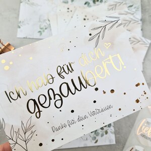 May include: A white card with gold foil lettering that reads 'Ich hab für dich gesagt!' and 'Danke für dein Vertrauen.' The card has a watercolor background with gold glitter and a small black and white illustration of a branch with leaves.