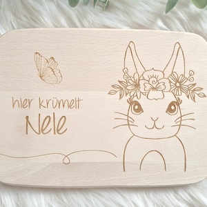 May include: A wooden cutting board with a bunny rabbit design and the text "hier krümelt  Nele". The bunny has a flower crown and a butterfly is in the top left corner.