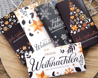 Pode incluir: Várias barras de chocolate com designs festivos de Natal. As embalagens apresentam flocos de neve, estrelas e elementos botânicos dourados e brancos. O texto nas embalagens diz "Frohe Weihnachten" e "Und ein gutes neues Jahr."