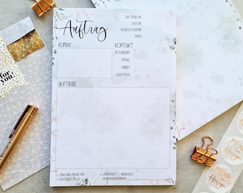 May include: A stationery set featuring a notepad with the word "Auftrag" and decorative floral accents. The set includes a pen, a gold-colored paper clip, and stickers with the text "fee Dich vom Herzen". The notepad has sections for customer details and order information.