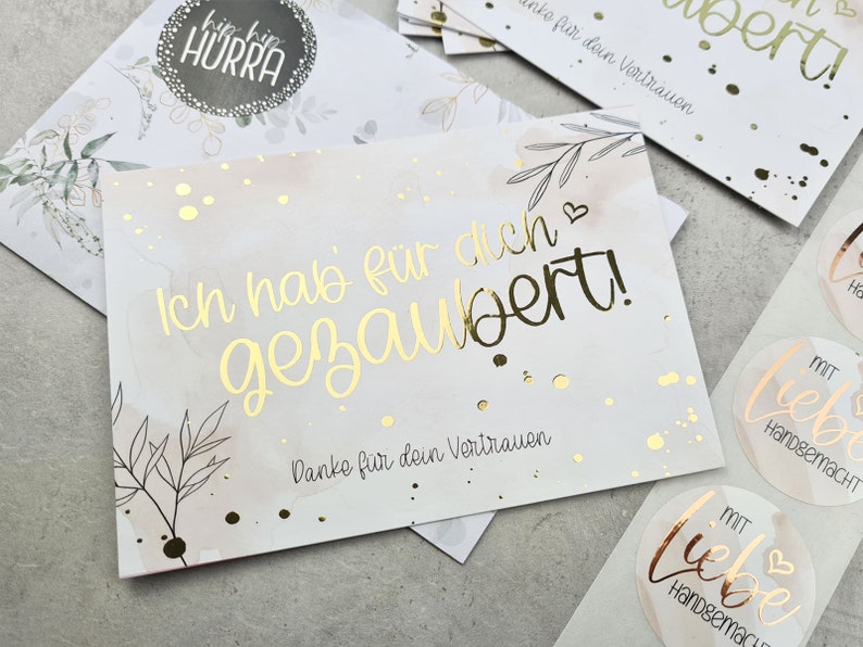 May include: A white card with gold lettering that reads 'Ich hab für dich gezaubert!' with a small heart symbol. The card has a watercolor background with gold glitter. The card also has the text 'Danke für dein Vertrauen' in gold lettering. The card is surrounded by other cards and stickers with gold lettering and watercolor backgrounds.
