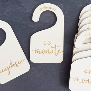Może przedstawiać: Zestaw drewnianych etykiet na wieszaki do ubrań dla niemowląt, z rozmiarami i przedziałami wiekowymi w języku niemieckim. Etykiety są w różnych rozmiarach, przy czym największa etykieta to "Größe 56-62 1-3 Monate", a najmniejsza to "Größe 50 neugeboren".
