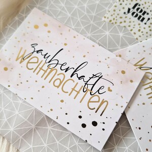 May include: Greeting cards with the German phrase "zauberhafte Weihnachten" in gold lettering, meaning "magical Christmas." The cards have a watercolor-style pink and white background with gold speckles. A smaller card with the text "for you!" is also visible.