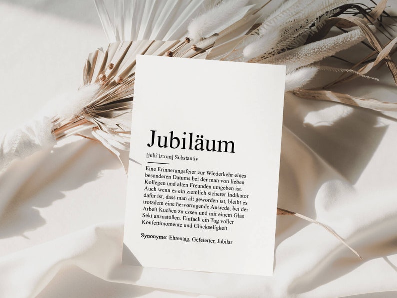May include: A white card with black text defining the German word "Jubil&auml;um" as a noun. The definition explains that it is a celebration of a special date, often with colleagues and friends. It also mentions that it is a reminder of getting older, but still a good excuse to have cake and champagne. The card is on a white surface with a dried palm leaf.