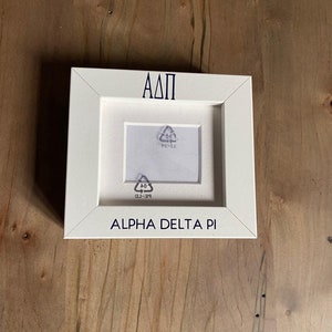 May include: White picture frame with a small opening for a photo. The frame has the letters "ΑΔΠ" and the words "Alpha Delta Pi" printed in black on the front.