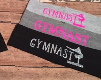 May include: Two gray and black headbands with the word "Gymnast" written in different colors and fonts. One headband has the word "Gymnast" in red, blue, and silver glitter. The other headband has the word "Gymnast" in pink and silver glitter.