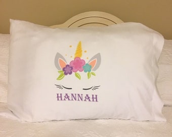 May include: White pillowcase with a unicorn face and flowers embroidered on it. The unicorn has a gold horn and closed eyes. The name "HANNAH" is embroidered below the unicorn.