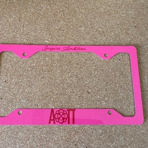 May include: Pink plastic license plate frame with the text "Inspire Ambition" and the letters "AOT" with a flower design in the center.