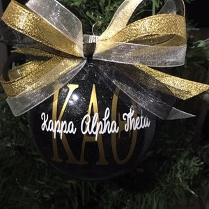 May include: Black Christmas bauble with gold glitter and white lettering that reads "Kappa Alpha Theta" and "KAO". The bauble is decorated with a black, gold, and white ribbon bow.