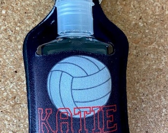 Op de afbeelding: Zwarte handdesinfectiehouder met een witte volleybalafbeelding en de naam "KATIE" in rode tekst. De houder heeft een zilveren ring om aan een sleutelhanger te bevestigen.