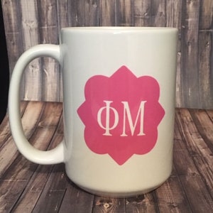 May include: White ceramic mug with a pink monogram of the letters "ΦM" in a decorative flower shape.