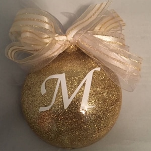 May include: Gold glitter Christmas ornament with a white letter M on it. The ornament is decorated with a white and gold ribbon.