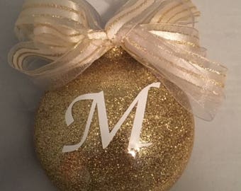 May include: Gold glitter Christmas ornament with a white letter M on it. The ornament is decorated with a white and gold ribbon.