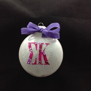 May include: A white glitter Christmas ornament with a purple ribbon and a pink and purple "ΣΚ" monogram.