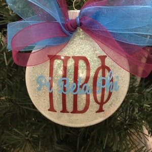 May include: A white glitter Christmas ornament with a red and blue ribbon bow. The ornament has the letters "ΠΒΦ" and the words "Pi Beta Phi" written on it.