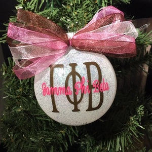 Peut inclure: Boule de Noël blanche à paillettes avec un nœud en ruban brun et rose. La boule est ornée des lettres "ΓΦΒ" en or et du texte "Gamma Phi Beta" en rose.