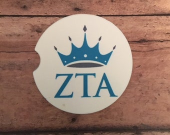 May include: White car coaster featuring a blue crown design and the letters "ZTA" in blue. The coaster has a small notch for easy removal from a car cup holder. The design is centered on the coaster.