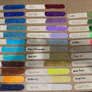 Op de afbeelding: Een verzameling houten knutselstokjes, elk met een andere glitterkleur. De kleuren zijn onder meer blauw, paars, groen, goud en roze, met handgeschreven labels zoals "Blue Jean", "Rood" en "Lime".
