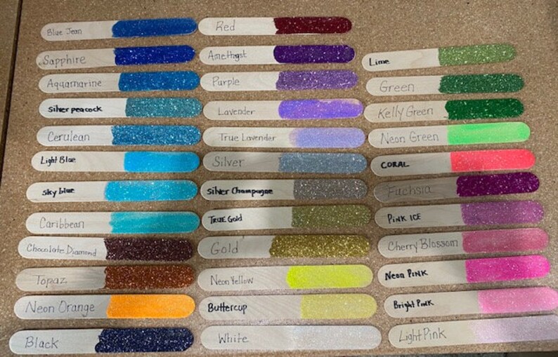 Op de afbeelding: Een verzameling houten knutselstokjes, elk met een andere glitterkleur. De kleuren zijn onder meer blauw, paars, groen, goud en roze, met handgeschreven labels zoals "Blue Jean", "Rood" en "Lime".