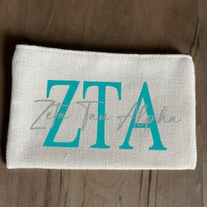 May include: A white canvas zippered pouch with the letters "ZTA" in teal blue. The words "Zeta Tau Alpha" are written in a smaller font below the letters.