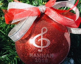 Könnte beinhalten: Roter Glitzer-Weihnachtsbaumschmuck mit einem silbernen Violinschlüssel und dem Text "HANNAH 2018" darauf. Der Schmuck ist mit einem roten und silbernen Band verziert.