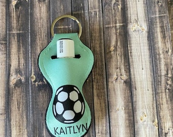Op de afbeelding: Een turquoise blauwe stoffen sleutelhanger met een wit voetbalontwerp en de naam "KAITLYN" gedrukt op de onderkant. De sleutelhanger heeft een metalen ring en een transparante plastic buis voor het bewaren van lippenbalsem of handdesinfectie.