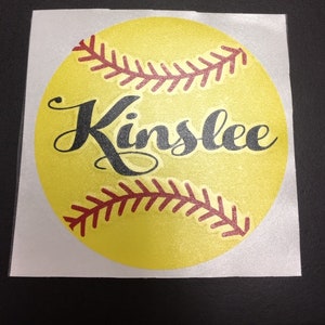 May include: A yellow softball with red stitching and the name "Kinslee" written in black cursive.