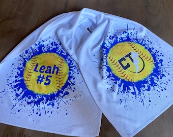 May include: White softball towel with blue paint splatter design. One side features a yellow softball with "Leah #5" printed on it. The other side features a yellow softball with "CT" printed on it.