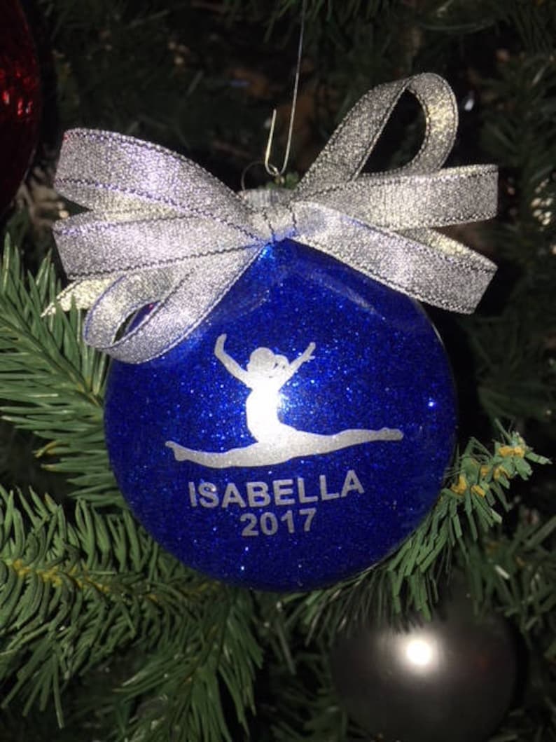 Op de afbeelding: Een blauwe glitter kerstbal met een zilveren strik. De kerstbal heeft een turnster silhouet en de naam "ISABELLA" met het jaar "2017" in witte letters. De kerstbal hangt aan een kerstboom.