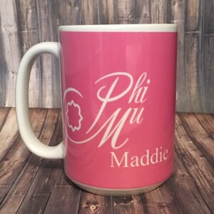 May include: A pink ceramic mug with a white design that says "Phi Mu Maddie".