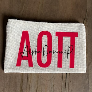 May include: A white canvas zippered pouch with the letters AOT in red and the text "Alpha Omicron Pi" in black cursive.