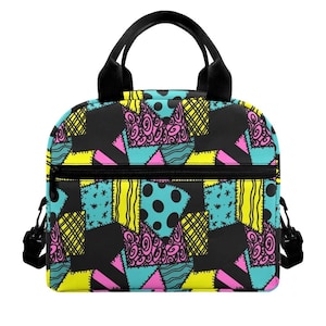 May include: A lunch bag featuring a patchwork pattern of black, bright yellow, turquoise, and pink fabric segments. The design includes various geometric shapes, polka dots, swirls, and cross-hatch patterns. It has black handles and a front zipper.