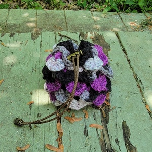 May include: A handmade crochet drawstring bag in shades of black, purple, gray, and white. The bag features a textured, scaly pattern and a brown leather drawstring with a decorative metal end. The bag is sitting on a weathered green wooden surface.