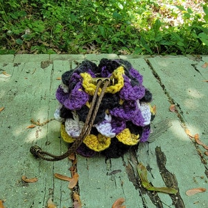 May include: A crocheted dice bag with a purple, yellow, black, and white color scheme. The bag has a drawstring closure and is sitting on a weathered wooden surface.