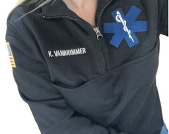 Op de afbeelding: Zwarte trui met een rits en een blauwe ster van levenspatch. De tekst "K. VANBRIMMER" is geborduurd op de voorkant van de trui.