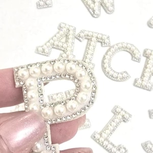 May include: A set of pearl and rhinestone letters, including the letters F, W, A, T, C, H, I, and P. The letters are white and have a shiny finish.