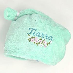 May include: A light green, absorbent hair towel with the name "Tiarra" embroidered in cursive script. The towel features a floral design in pink and white. The towel is designed to wrap around the hair and has a knotted top.