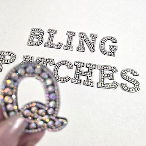 May include: Rhinestone letters spelling "BLING PATCHES" with a single letter "Q" in the foreground. The letters are silver with clear rhinestones.