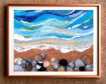 Op de afbeelding: Een ingelijst naaldviltwerk dat een zeelandschap voorstelt met blauwe en groene golven die breken op een zandstrand met bruine en grijze stenen.