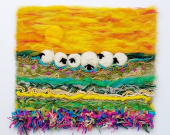 Op de afbeelding: Een kleurrijk wollen wandkleed dat een zonsondergangscène voorstelt met een rij witte schapen voor een gele en oranje lucht. De schapen zijn zwart omlijnd en omgeven door groene en blauwe wol.