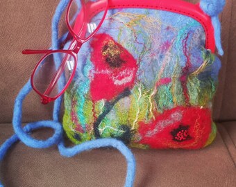May include: A small blue felt purse with a red clasp and a red felt floral design. The purse has a blue strap and is sitting on a brown surface.