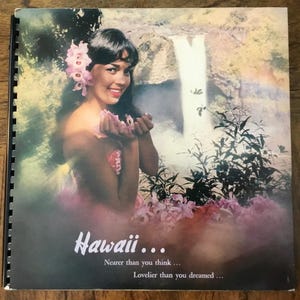 May include: A vintage travel brochure featuring a woman with pink flowers in her hair, smiling and holding out her hand in front of a waterfall. The text on the brochure reads "Hawaii... Nearer than you think... Lovelier than you dreamed..."