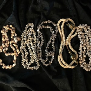 May include: Five necklaces made of small, natural seashells in varying shades of beige, brown, and gray. The necklaces are arranged on a dark, textured fabric, showcasing their unique textures and shapes. The necklaces are a mix of different styles.