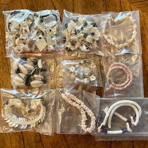May include: A collection of shell necklaces and bracelets in clear plastic bags. The jewelry features various shell shapes and colors, including white, black, and pink. Some pieces are strung with beads, and the arrangements vary in style and size.